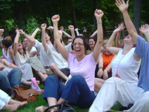 Lachende Menschen der europäischen Lachyoga Gruppe, sitzen zusammen und strecken die Arme gen Himmel.