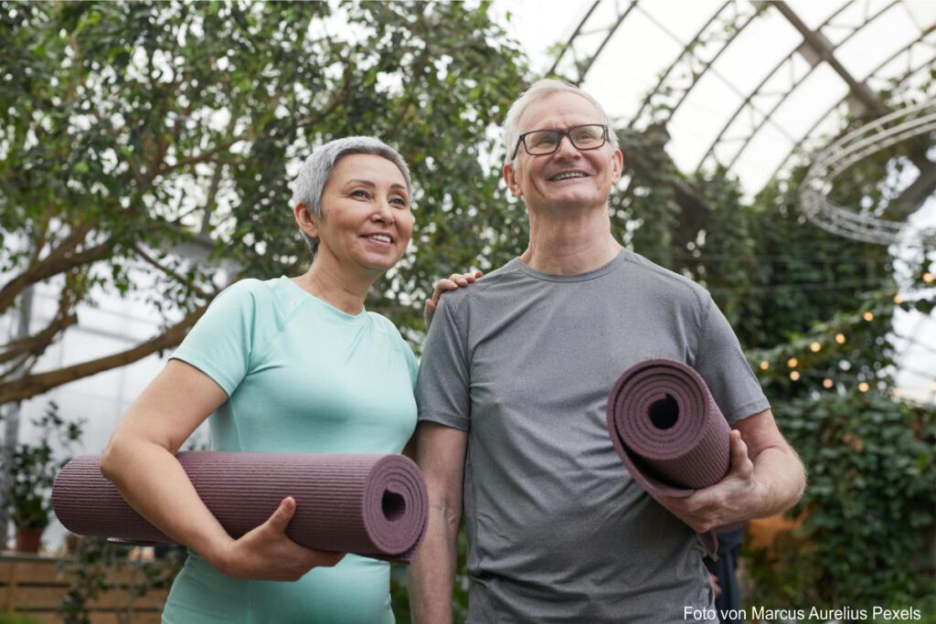 Eine Frau und ein Mann älteren Alters stehen mit zusammen und haben jeweils eine Yogamatte unterm Arm.